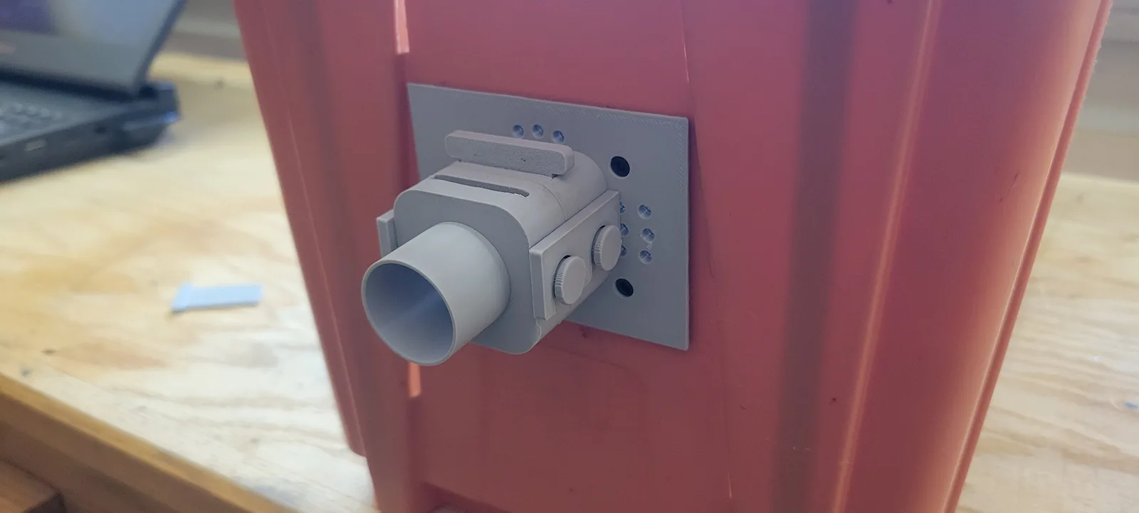 Close up of the tunnel adapter fitted to the outside end wall of an orange nuc box: a grey printed base plate with a cluster of small vent holes, a raised landing bar above the opening, and a quick connect body carrying a 1 inch hose socket held on by two knurled thumb knobs.