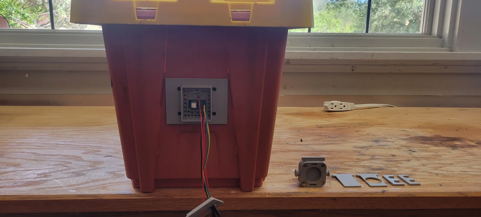 The end wall of an orange nuc box on a workbench by a window, with a grey printed rear plate fitted. A square recessed pocket in the plate is perforated with a dense grid of small vent holes, and a small square CO2 sensor module sits in the centre with four coloured wires running out of it and down the side of the box.