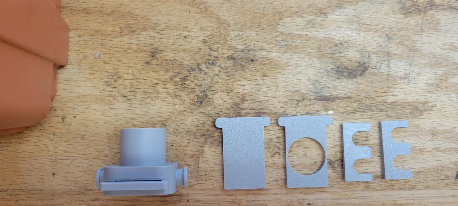 Grey 3D printed PETG parts laid out on a wooden workbench beside the corner of an orange plastic nuc box: a quick connect body with a cylindrical hose socket and two thumb knobs, two flat T-shaped doors, one solid and one with a large circular opening, and two small C-shaped latch clips.